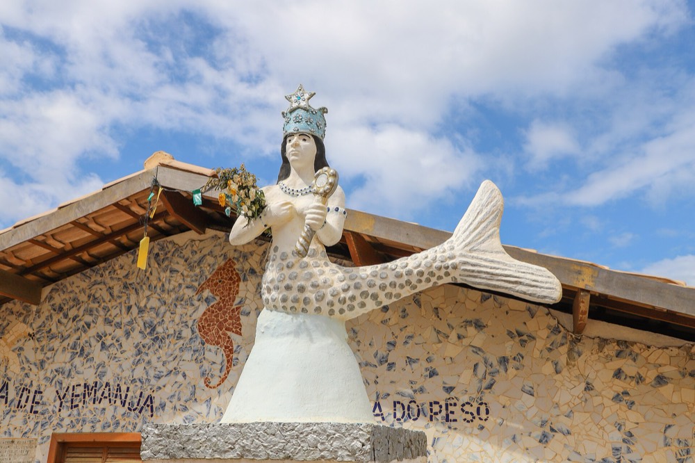 Imagem de Yemanjá como sereia coroada, segurando flores, em frente a uma parede de mosaico